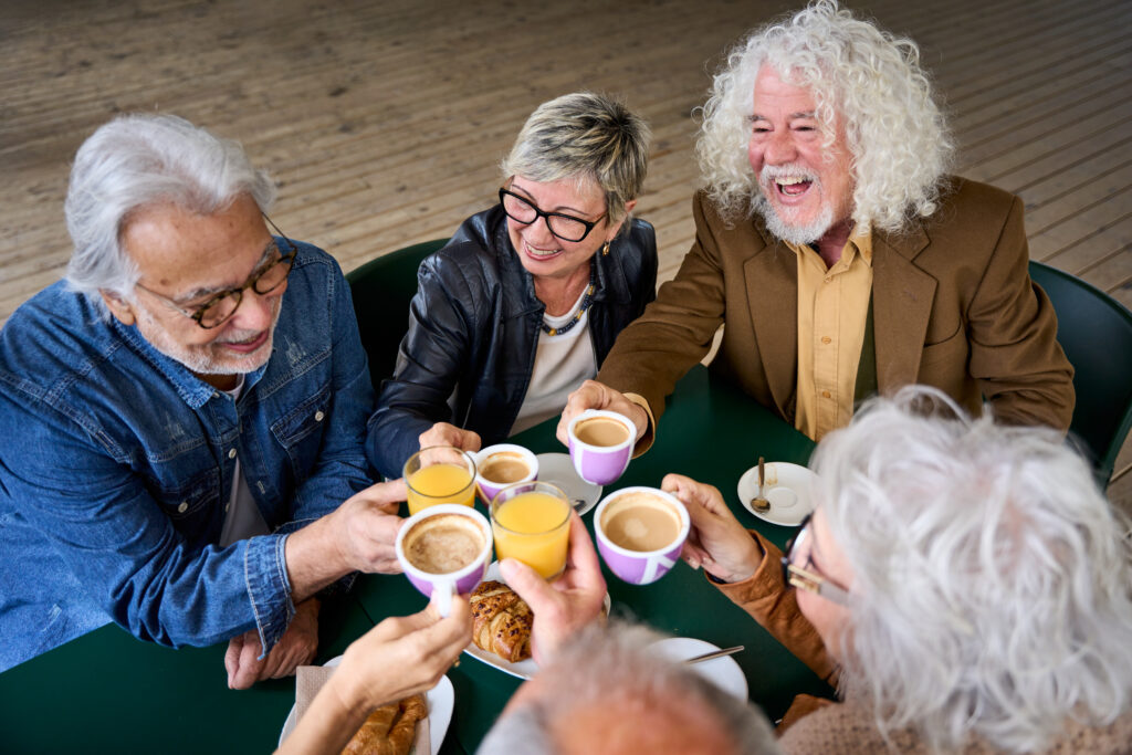 A group of six older adults sits around a table at the lakeside ranch, smiling and raising cups in a toast. They appear to be enjoying coffee and pastries together, reveling in the relaxing park activities they’ve experienced earlier.